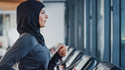 Obraz na płótnie Canvas Athletic Muslim Sports Woman Wearing Hijab and Sportswear Running on Treadmill. Energetic Fit Female Athlete Training in Gym Alone. Urban Business District Window View. Side View Portrait