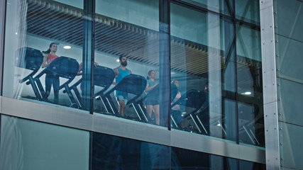 Athletic People Running on Treadmills, Doing Fitness Exercise. Fit and Muscular Athletes Actively Training in the Modern Gym. Sports People Workout. Low Angle Shot From Outside the Building
