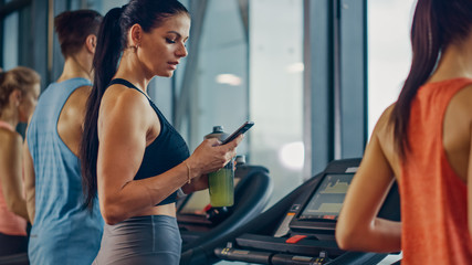 Fototapeta na wymiar Beautiful Athletic Sports Woman in Gym Running on a Treadmill, Uses Smartphone and Drinks Protein Supplement Hydration Liquid From Tumbler. In Background Fit Athletes Training. Side View