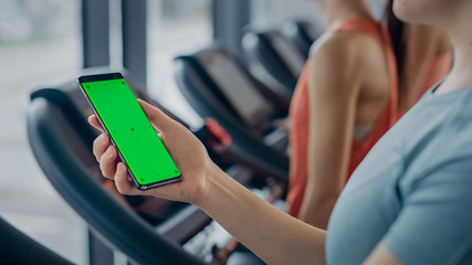 Athletic Woman Uses Green Mock-up Screen Smartphone while Running on the Treadmill in a Gym. Sports Female Posts on Social Media, Takes Pictures, Watches Videos while Exercising in the Fitness Club