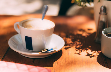 Close-up white cup with hot latte with spoon on a brown wooden table in a street cafe, with blurred background. Soft focused mug of coffee outdoor. Paper shaker and a part of light pot with flowers