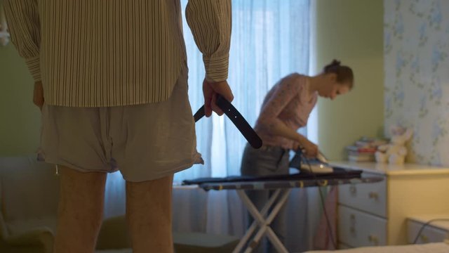 A man in shorts and a shirt holding a belt in his hands waiting while his wife ironing trousers for him. Dominant aggressive husband, domestic violence