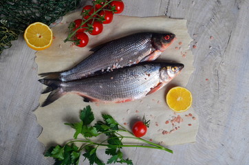 Two fresh fish vimba on baking sheet, gray wooden background, decorated with green fishing net, small tomatoes, parsley and lemon