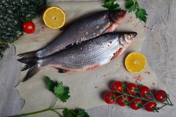 Two fresh fish vimba on baking sheet, gray wooden background, decorated with green fishing net, small tomatoes, parsley and lemon