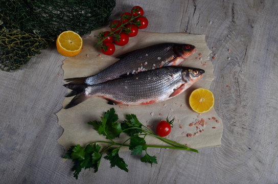 Two fresh fish vimba on baking sheet, gray wooden background, decorated with green fishing net, small tomatoes, parsley and lemon
