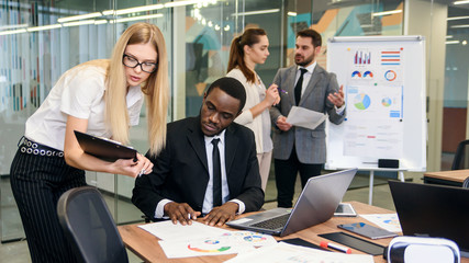 Blond businesswoman in stylish glasses gives for signature different documents to purposeful dark-skinned manager in the meeting room with other team mates at the background.