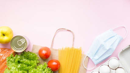 Coronavirus special food supplies background concept. Bag with medical protective mask, tomatoes, salad, eggs, apple, canned food on pink background. Top view. Copy space. 