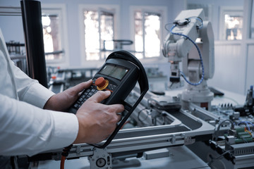 Close up of man's hand holding teach pendant to control robot arm which is integrated on smart factory production line. industry 4.0 automation line equipped with sensors and robot at background.