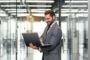 Fototapeta premium Smiling bearded business man focused on company datas while standing in office corridor and using laptop.