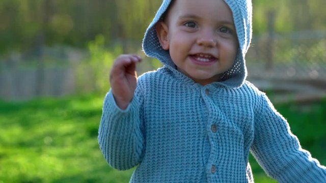 Cute portrait of walking smiling little baby boy waving hello in park or green garden. First steps. Lovely son, family, toddler concept.