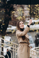 Outdoor photo of brunette lady posing with black umbrella in rainy autumn day.Fashion street style portrait. wearing dark casual trousers, white sweater and creamy coat.Fashion concept.