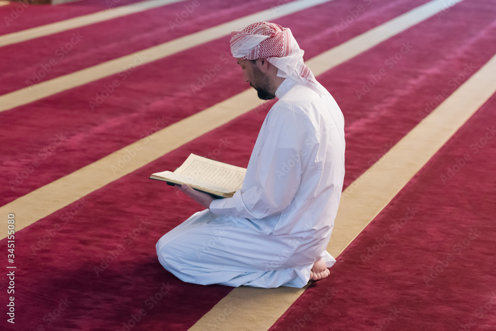 Wall mural Young Arabic Muslim man reading Koran and praying. Religious muslim man reading holy koran inside the mosque.