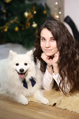 Portrait of a woman with dark hair, she is lying on the floor with a white dog, against the background of a Christmas tree