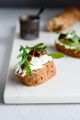 Italian bruschetta (sandwich, crostini, toast) with confit tomatoes, feta, olive oil and arugula leaves on white marble background. Traditional italian appetizer or snack, antipasto. Selective focus