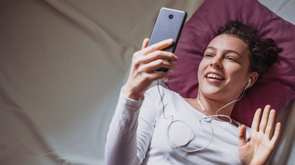 Young happy woman using mobile phone for video call , gesturing hi to friends, relatives or parents. Beautiful girl making facetime video calling with smartphone lying on the bed.