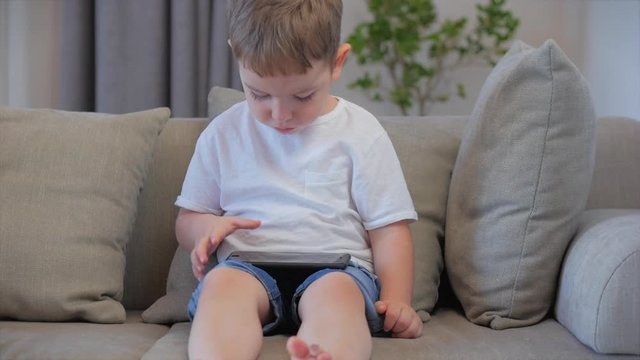 Curious cute boy preschool child using a digital tablet technology device looks telephone.Plays in the phone sitting on the sofa alone.Children tech addiction concept play game at home.