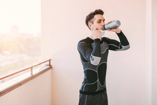 Exhausted thirsty sports man in protective medical face mask drinking water after exercising on balcony. Young tired man fitness coach holding water bottle in hand. Home workout, COVID-19 quarantine - Powered by Adobe