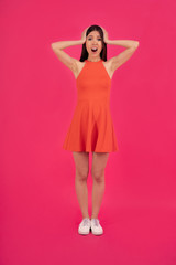 Image of joyous young asian woman expressing shocked expression and holding head by her hands isolated over pink background. dressed in orange dress