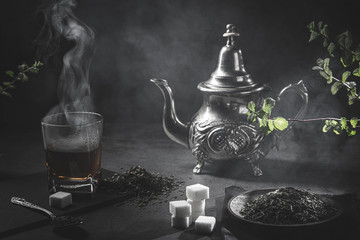 Traditional Moroccan teapot, with a steaming cup of tea, sugar and mint, in a smoky black...
