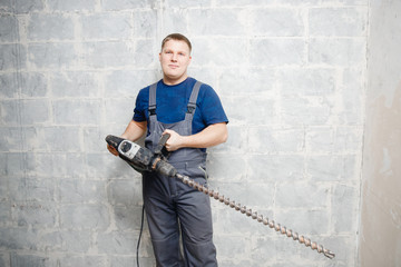 Male worker builder holds big drill background gray wall