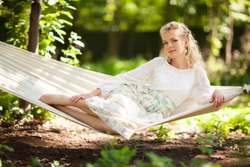 Front view of beautiful young woman resting on hanging hammock swing bed in spring forest. Charming lady in elegant long dress looking at camera. Concept of beauty, elegance and nature.