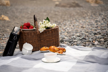 food, holidays and celebration concept - close picnic basket with thermos with tea, rolls, strawberries, flowers on the beach