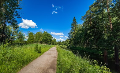 Summer landscape with a river, a path along the canal with water, trees, grass and a blue sky with white clouds in the Park