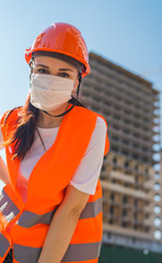 Portrait of female construction worker in medical mask and overalls on background of house under construction.
