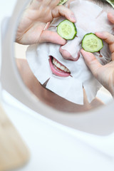 Caucasian cheerful man with a cosmetic mask on his face, holds slices of cucumber in front of his eyes. In the bathroom, smiling, reflection in the mirror. Home spa, facial skin care. Vertical shot.