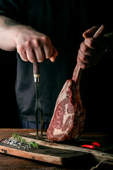 The chef's hands hold a raw Tomahawk a beef steak and a meat fork over a chopping Board