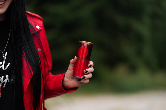 Woman In Red Leather Jacket Drinking Cool Soda Cola Drink From Red Can In Hand. Woman Drinking , Showing Blank Can. Girl Holding Energy Drink Or Other Drink. Caucasian Female Model On Background.