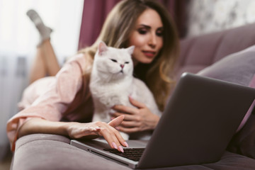 Fototapeta na wymiar A young pretty woman with a cute cat lies on a sofa and uses a laptop. The pet makes it difficult to work. Pestering cat. Self-isolation.