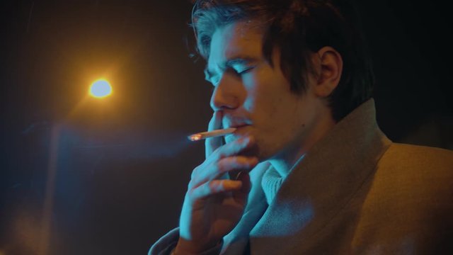 Young stylish man in white coat smoking cigarette outdoors