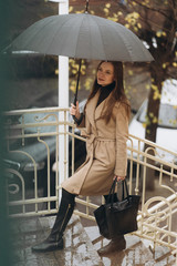 Outdoor photo of brunette lady posing with black umbrella in rainy autumn day.Fashion street style portrait. wearing dark casual trousers, white sweater and creamy coat.Fashion concept.