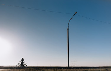 Silhouette of a young girl near the electric bicycle stand on the embankment on sunset background. Transportation in the village. Blue sky, streetlight and copy space.