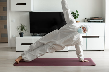 Young man in medical mask and in protective suit is practicing yoga on a mat at home. Stay home during COVID-19 quarantine concept. Yoga at home online. Coronavirus pandemic. Healthy lifestyle.