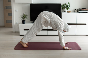 Young man in medical mask and in protective suit is practicing yoga on a mat at home. Stay home during COVID-19 quarantine concept. Yoga at home online. Coronavirus pandemic. Healthy lifestyle.