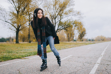 Fototapeta premium Young beautiful smiling girl in casual wear roller skating listens to music. Autumn walk in the fresh air.