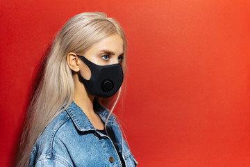 Obraz na płótnie Canvas Studio profile portrait of young blonde girl, wearing respiratory face mask of black color, against coronavirus. Background of red color with copy space.