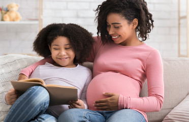 Cute Little Black Girl Reading Book With Pregnant Mom At Home