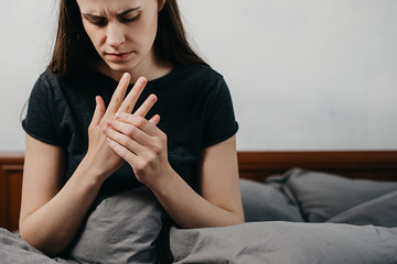 Sad tired young woman sitting on bed massaging hand suffering from rheumatoid arthritis concept,...
