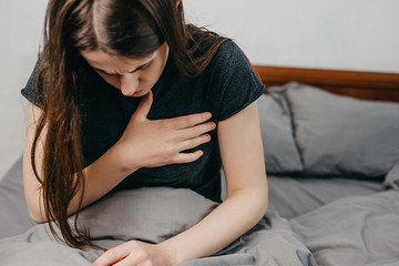 Front view of tired young woman feeling pain ache touching chest having heart attack. Sad worried girl suffers from heartache sitting on bed at home, infarction or female heart disease concept