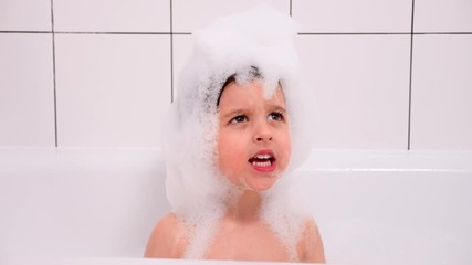three year old child with big brown eyes is sitting in a white bathroom with square tiles in foam and having fun