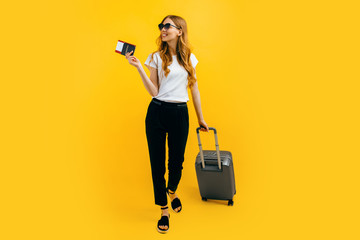 Happy beautiful woman, with a suitcase and passport with tickets on a yellow background. The concept of travel, business