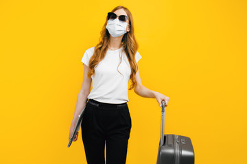 business woman with a protective medical mask on her face, with a laptop and a suitcase on a yellow background. The concept of travel, quarantine, and coronavirus