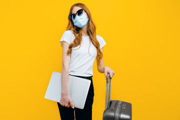 business woman with a protective medical mask on her face, with a laptop and a suitcase on a yellow background. The concept of travel, quarantine, and coronavirus