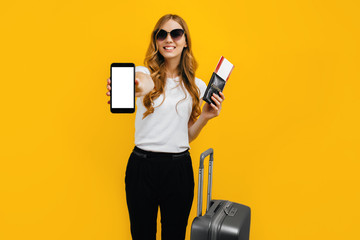 A happy woman in dark glasses, carrying a suitcase and a passport with tickets, shows an empty mobile phone screen for a copy of the space. The concept of travel, business