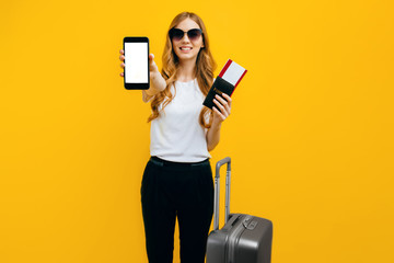 A happy woman in dark glasses, carrying a suitcase and a passport with tickets, shows an empty mobile phone screen for a copy of the space. The concept of travel, business