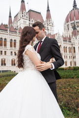 Fototapeta premium Bride and groom hugging in the old town street. Wedding couple walks in Budapest near Parliament House. Caucasian happy romantic young couple celebrating their marriage. Wedding and love concept.