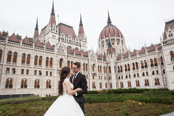 Bride and groom hugging in the old town street. Wedding couple walks in Budapest near Parliament House. Caucasian happy romantic young couple celebrating their marriage. Wedding and love concept.